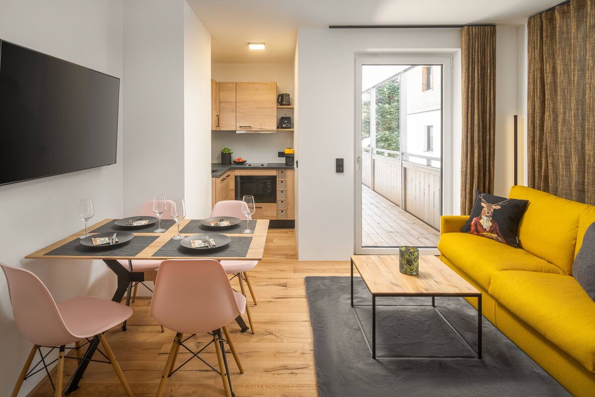 Una sala de estar moderna con un sofá amarillo, una mesa de comedor con sillas rosas y una cocina al fondo.