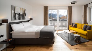 A modern hotel room with a large bed, a yellow sofa, a small table, and a view of mountains through the window. - Auhof (Bad Gastein)