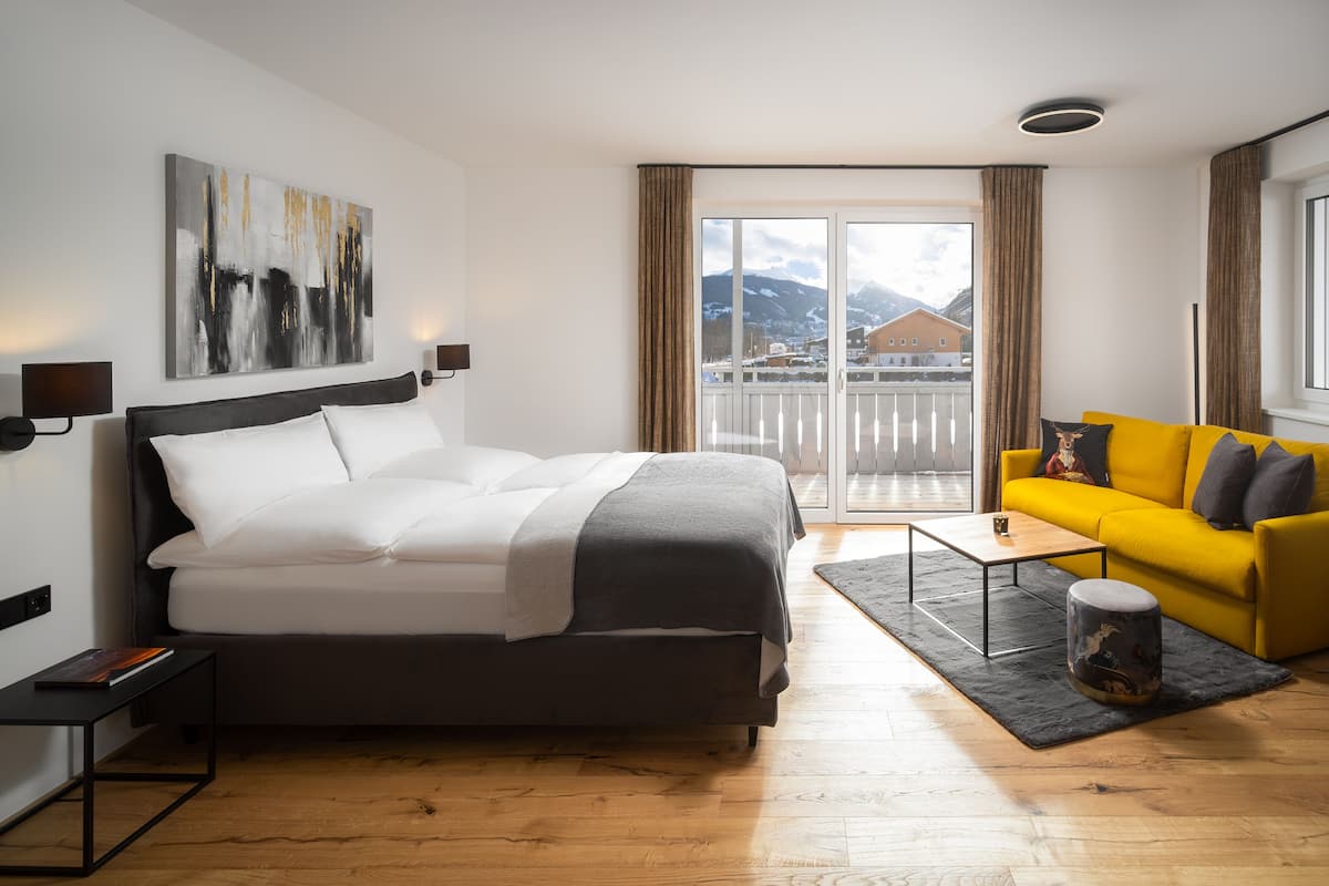 Habitación de hotel moderna con una cama grande, un sofá amarillo, una mesita y vistas a montañas a través del ventanal.