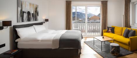 Habitación de hotel moderna con una cama grande, un sofá amarillo, una mesita y vistas a montañas a través del ventanal.