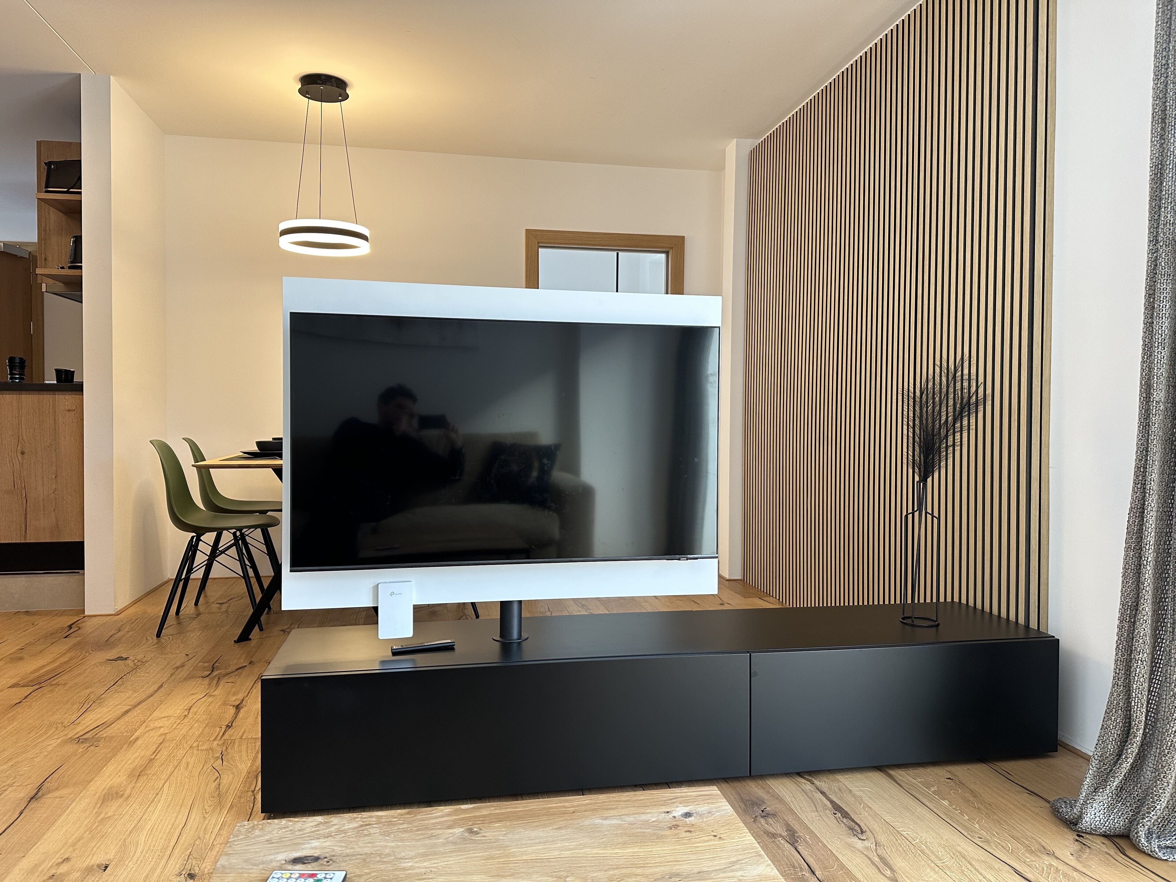 A modern living room with a large flat-screen TV on a black stand, a dining area with green chairs, and a wooden floor.