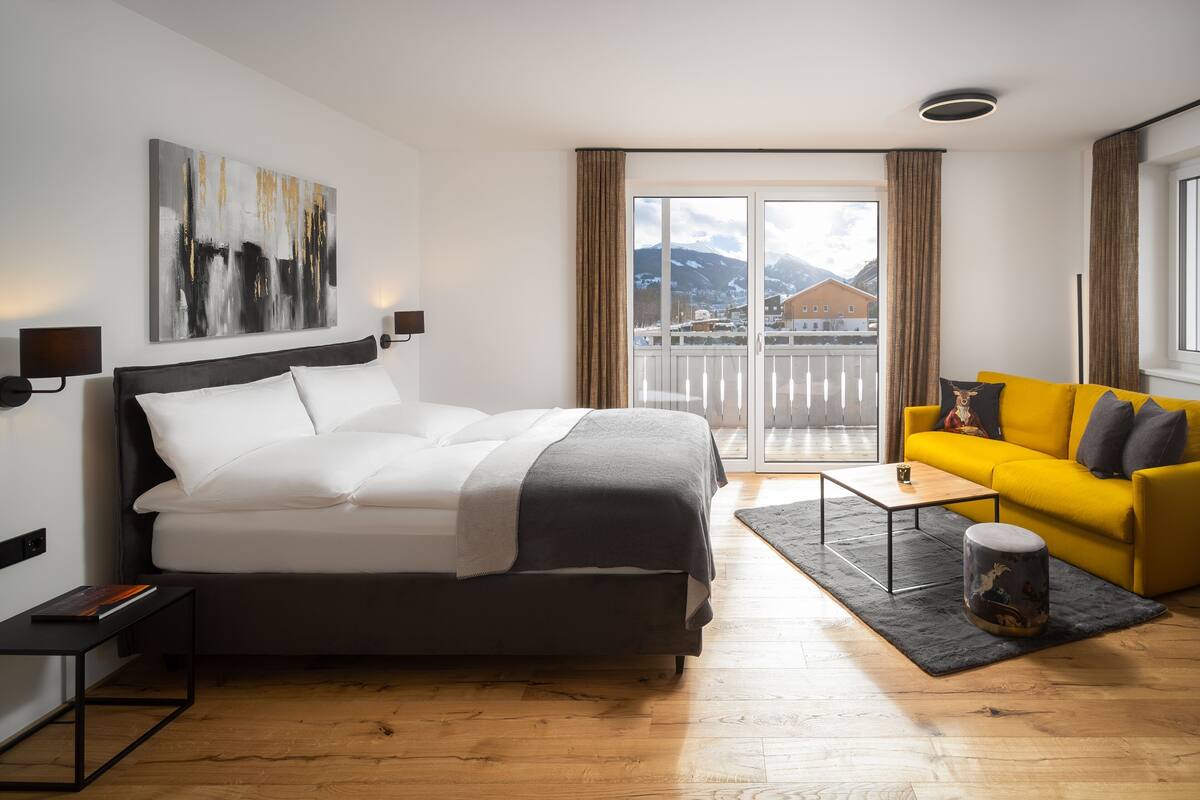 Habitación de hotel moderna con una cama, un sofá amarillo, una mesita y vistas a las montañas desde la ventana.