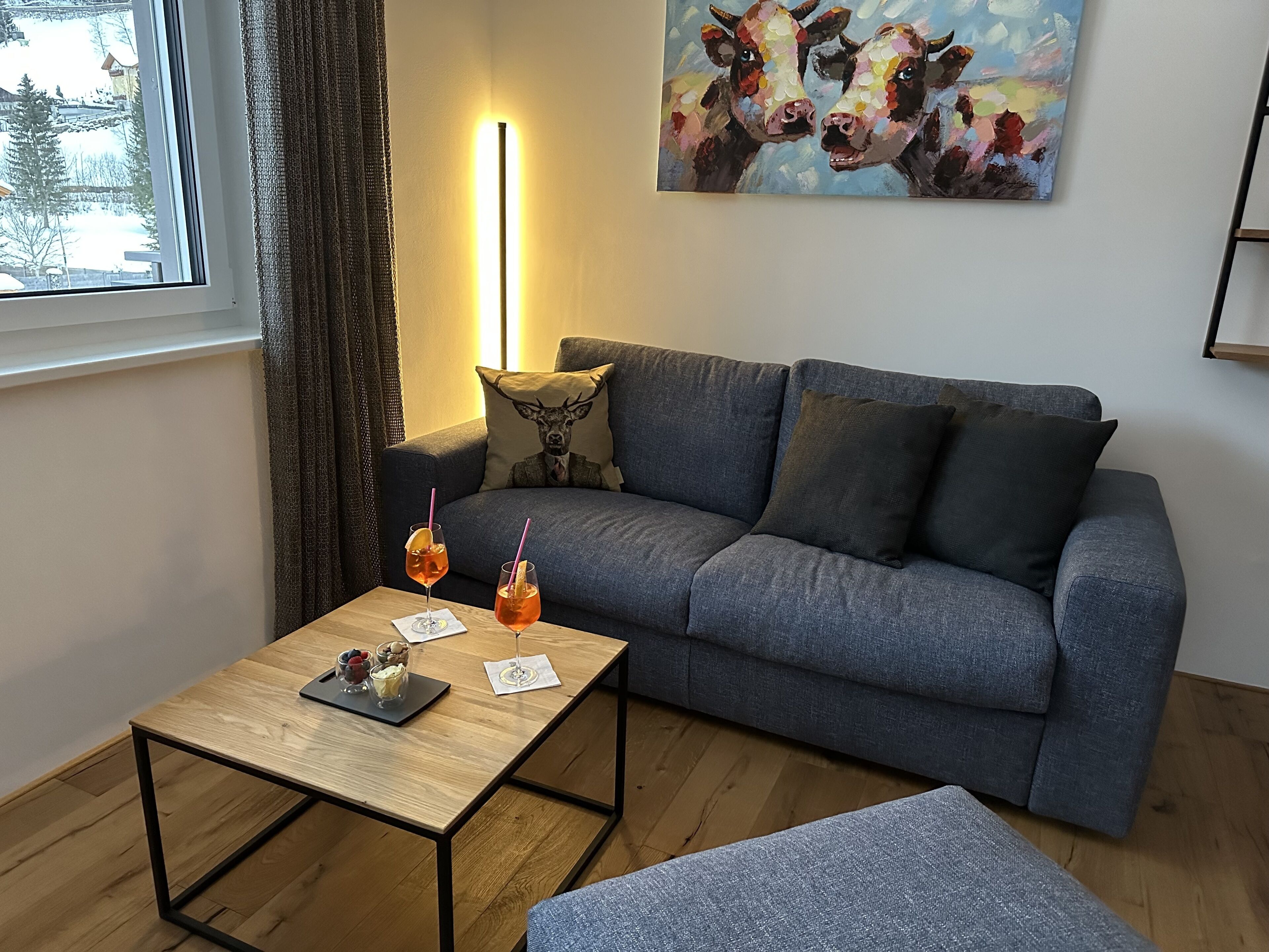 A living room with a grey sofa, a wooden coffee table with drinks, a painting of cows, and a window with curtains.