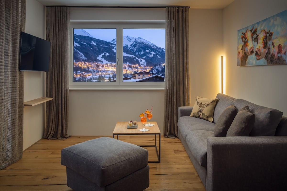 Una sala de estar moderna con un gran ventanal con vistas a un pueblo nevado, un sofá gris, una mesita con bebidas y un cuadro en la pared.