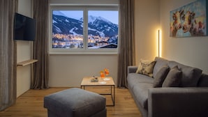 A modern living room with a large window overlooking a snowy mountain town, a grey sofa, a small table with drinks, and a painting on the wall. - Auhof (Bad Gastein)