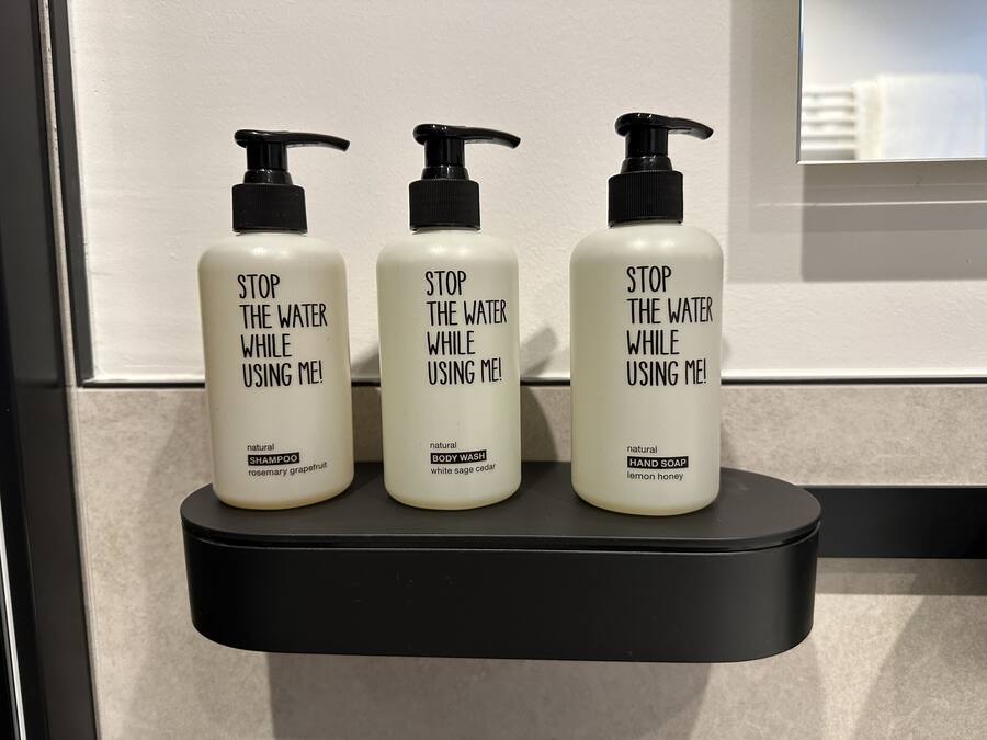Trois bouteilles de shampoing naturel, de gel douche et de savon pour les mains, posées sur une étagère noire.