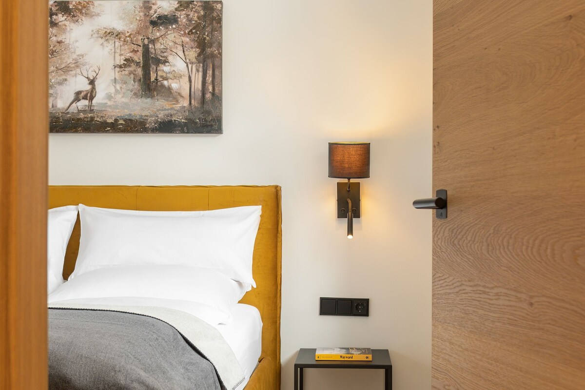 A bedroom with a bed, a nightstand with a lamp, a painting on the wall, and a wooden door.