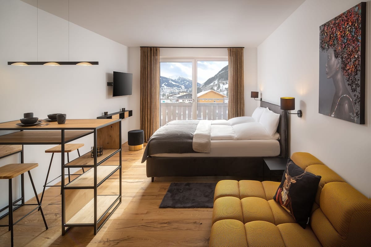 Una habitación de hotel moderna con cama, sofá, escritorio con taburetes, televisor y un gran ventanal con vistas a la montaña.