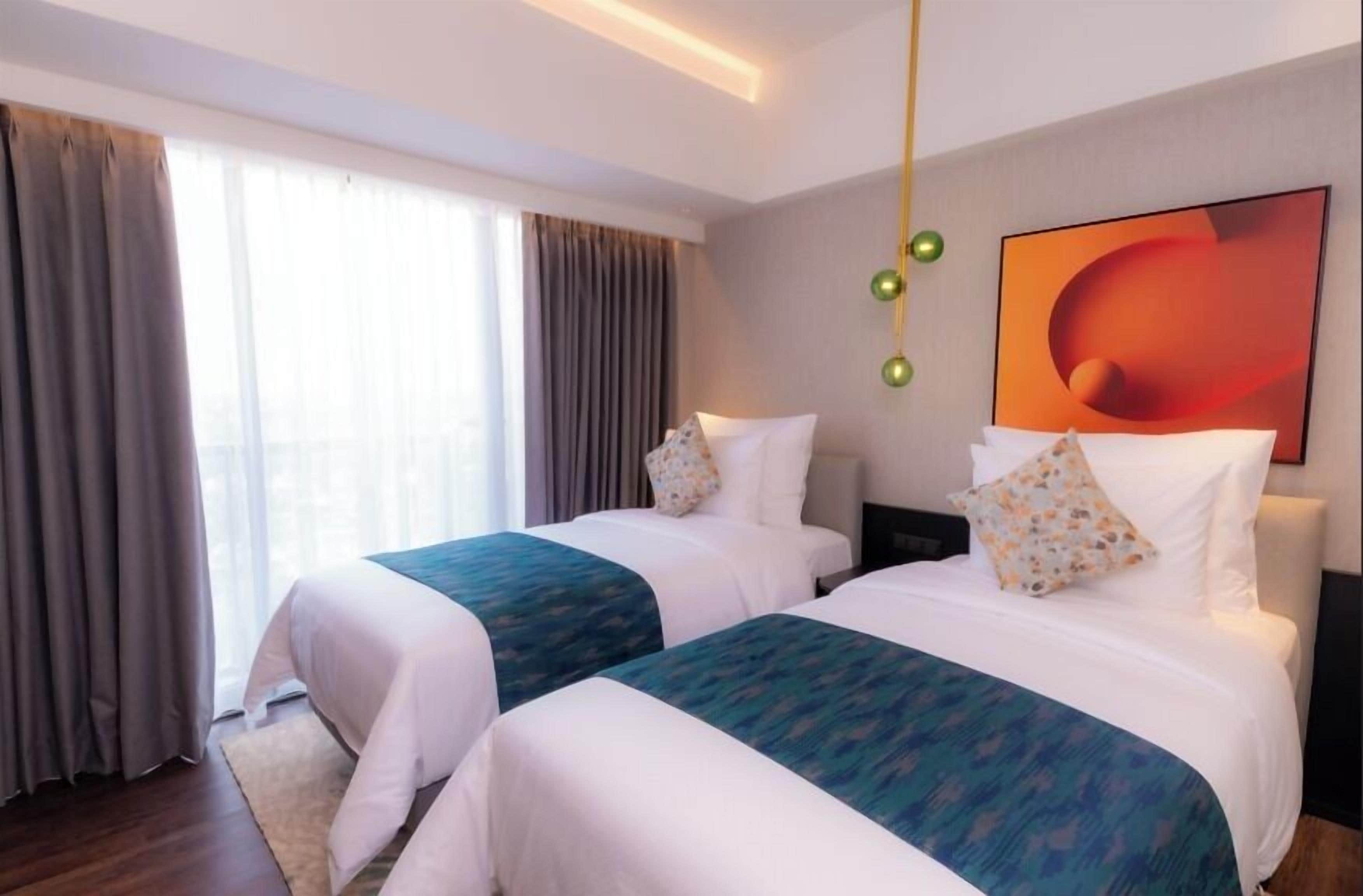 Une chambre d’hôtel avec deux lits, une grande fenêtre avec des rideaux, un tableau abstrait moderne et un luminaire suspendu.