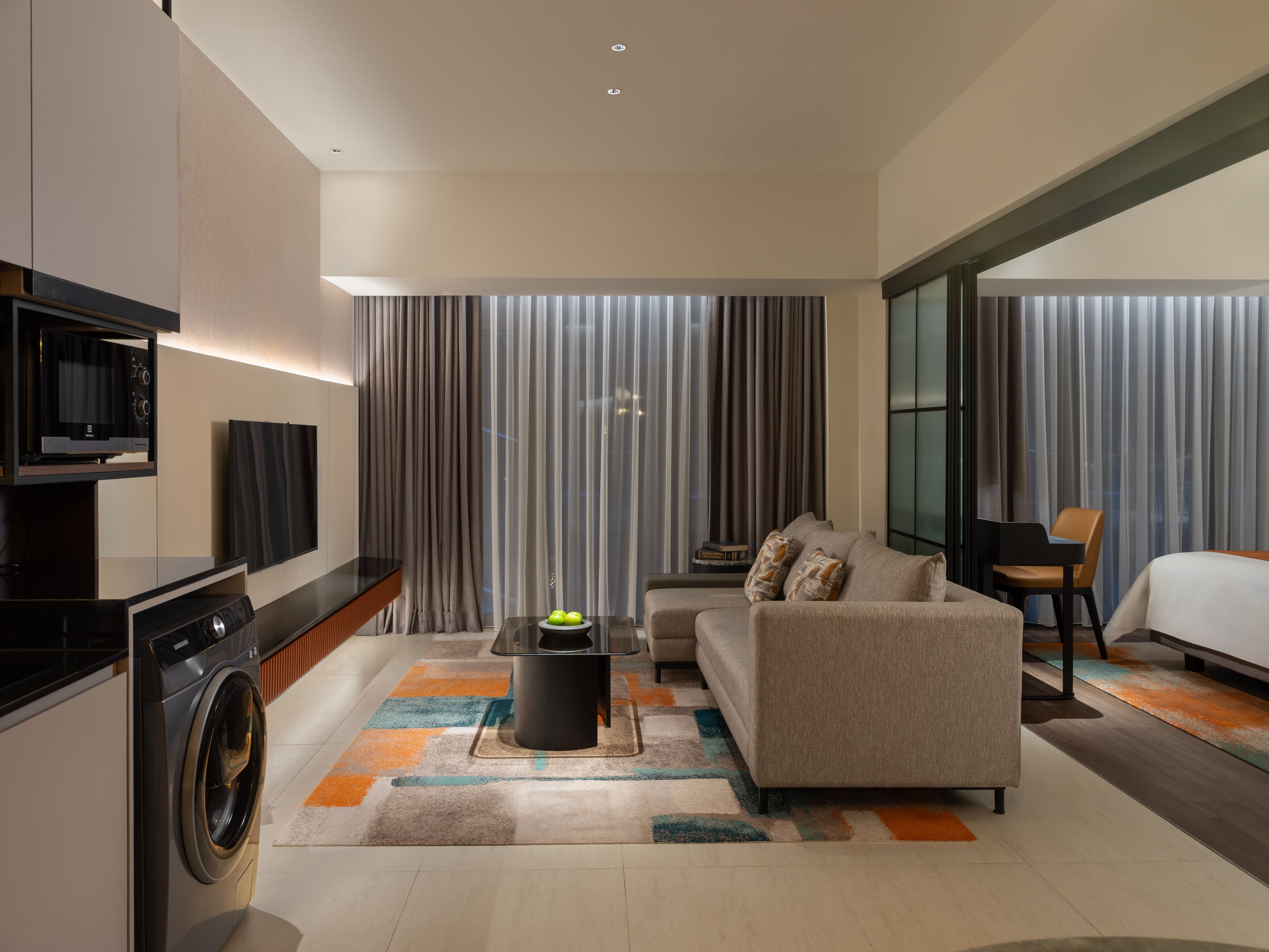 A modern hotel room with a sofa, a small table with a bowl of fruit, a washing machine, a flat-screen TV, and a large window with curtains.