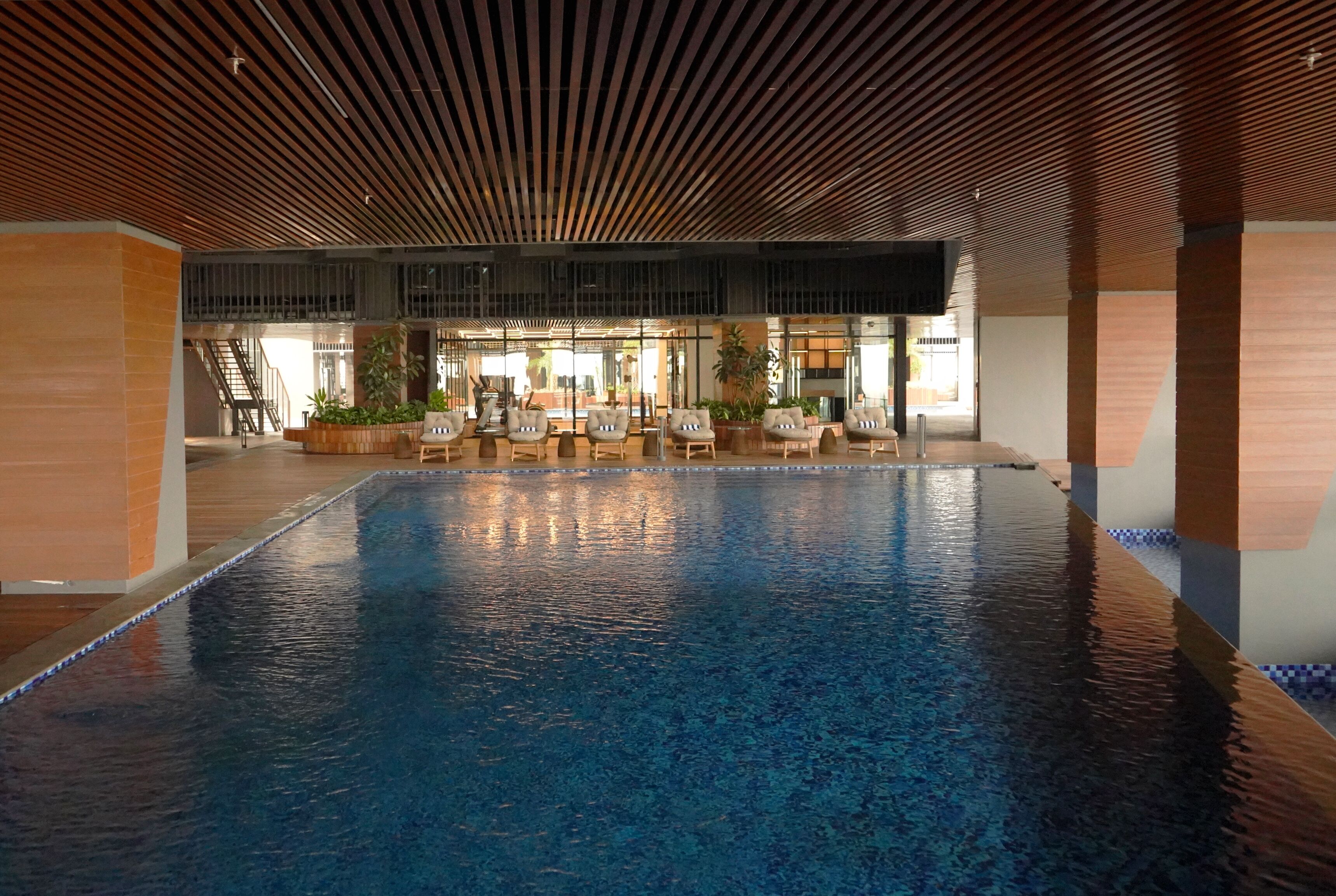 An indoor swimming pool with a wooden ceiling, seating area with chairs and potted plants, and a view of the outside.