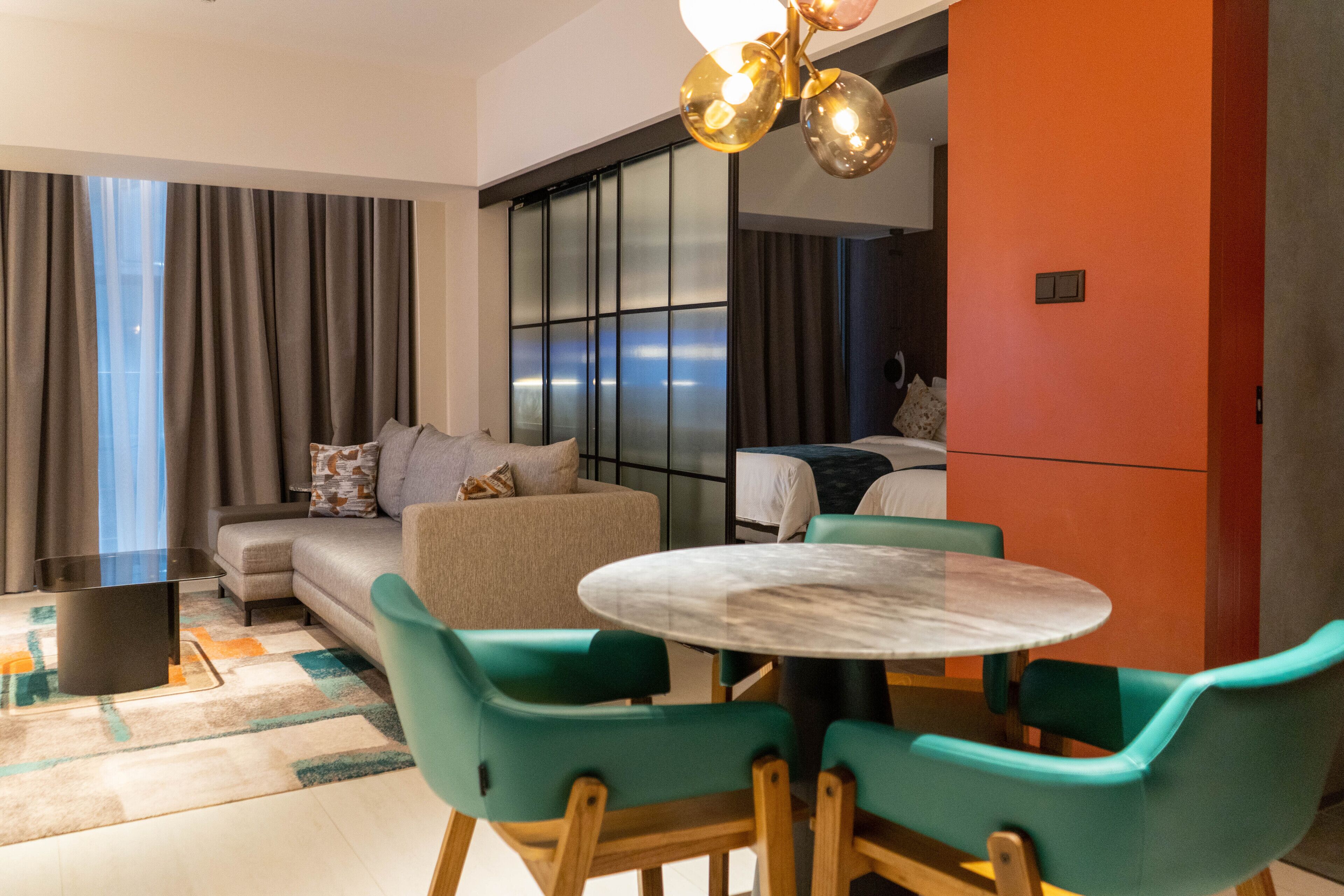 A modern hotel room with a sofa, a dining table with chairs, a large window with curtains, and a unique chandelier.