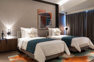 Habitación de hotel con dos camas, un cuadro abstracto grande en la pared, una mesita de noche con lámpara y vistas a la ciudad a través de la ventana.