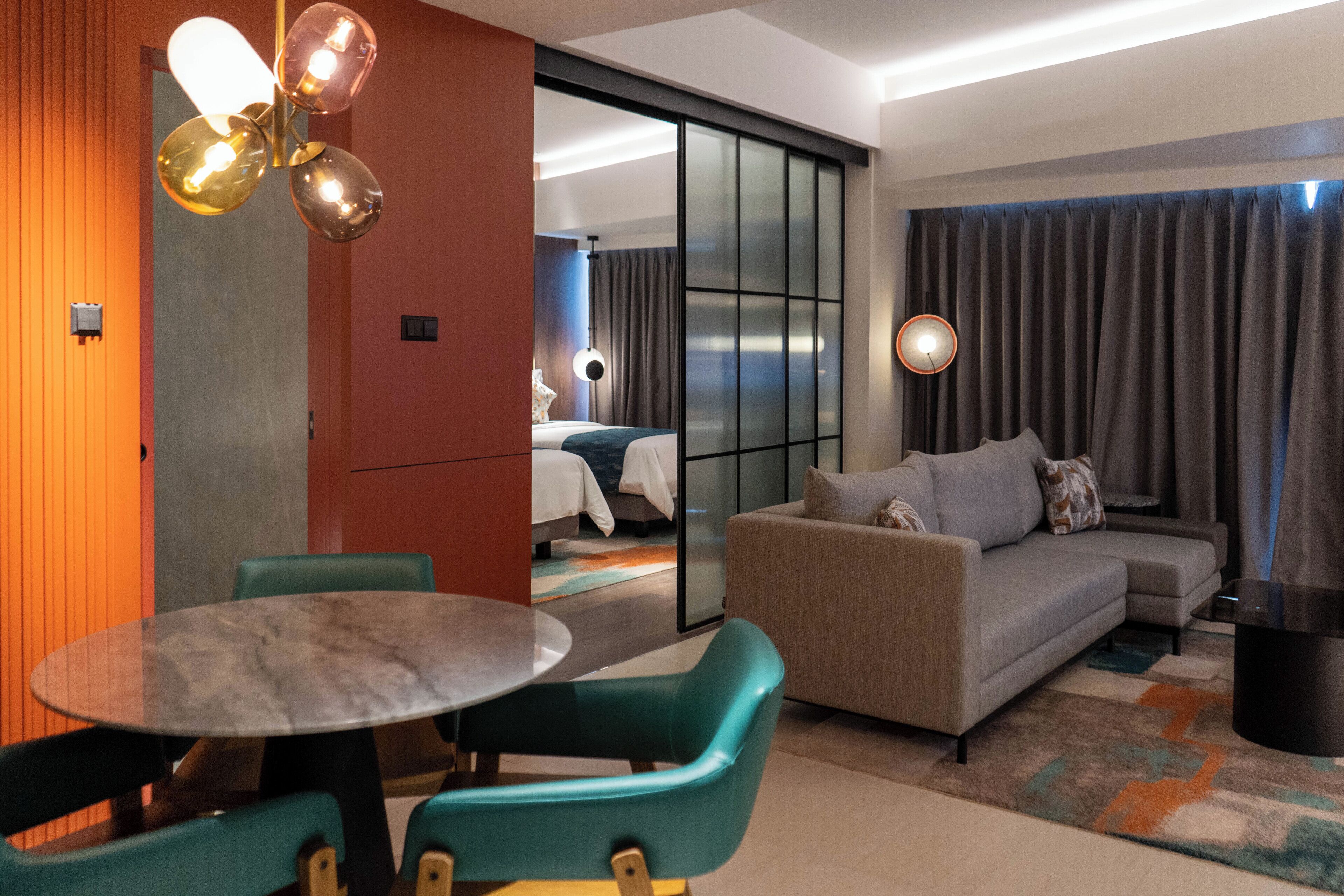 A modern hotel room with a sofa, a dining table, and chairs. There is a sliding door leading to another room.