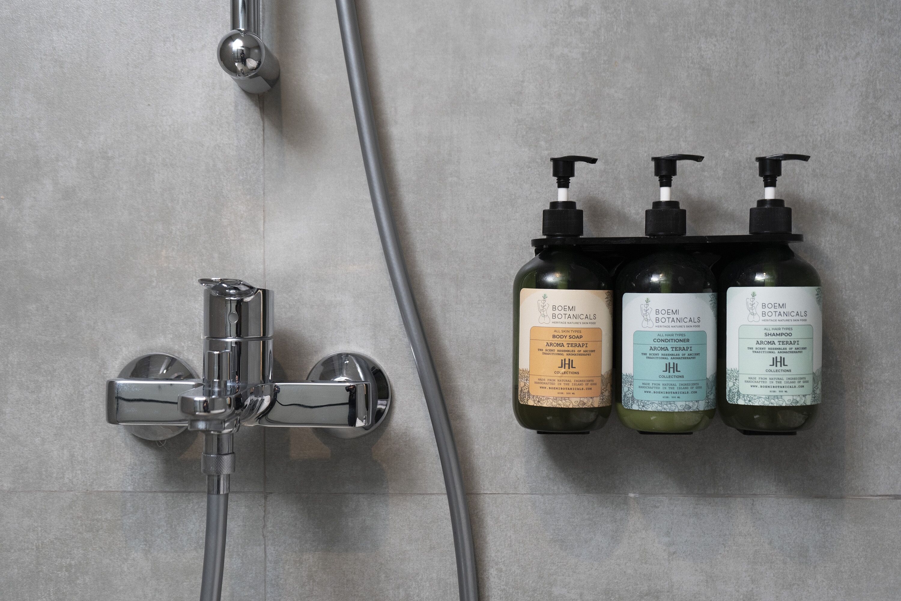 Área de chuveiro com torneira cromada e chuveiro manual. Três frascos de sabonete líquido, condicionador e shampoo Boemi Botanicals estão dispostos na parede.