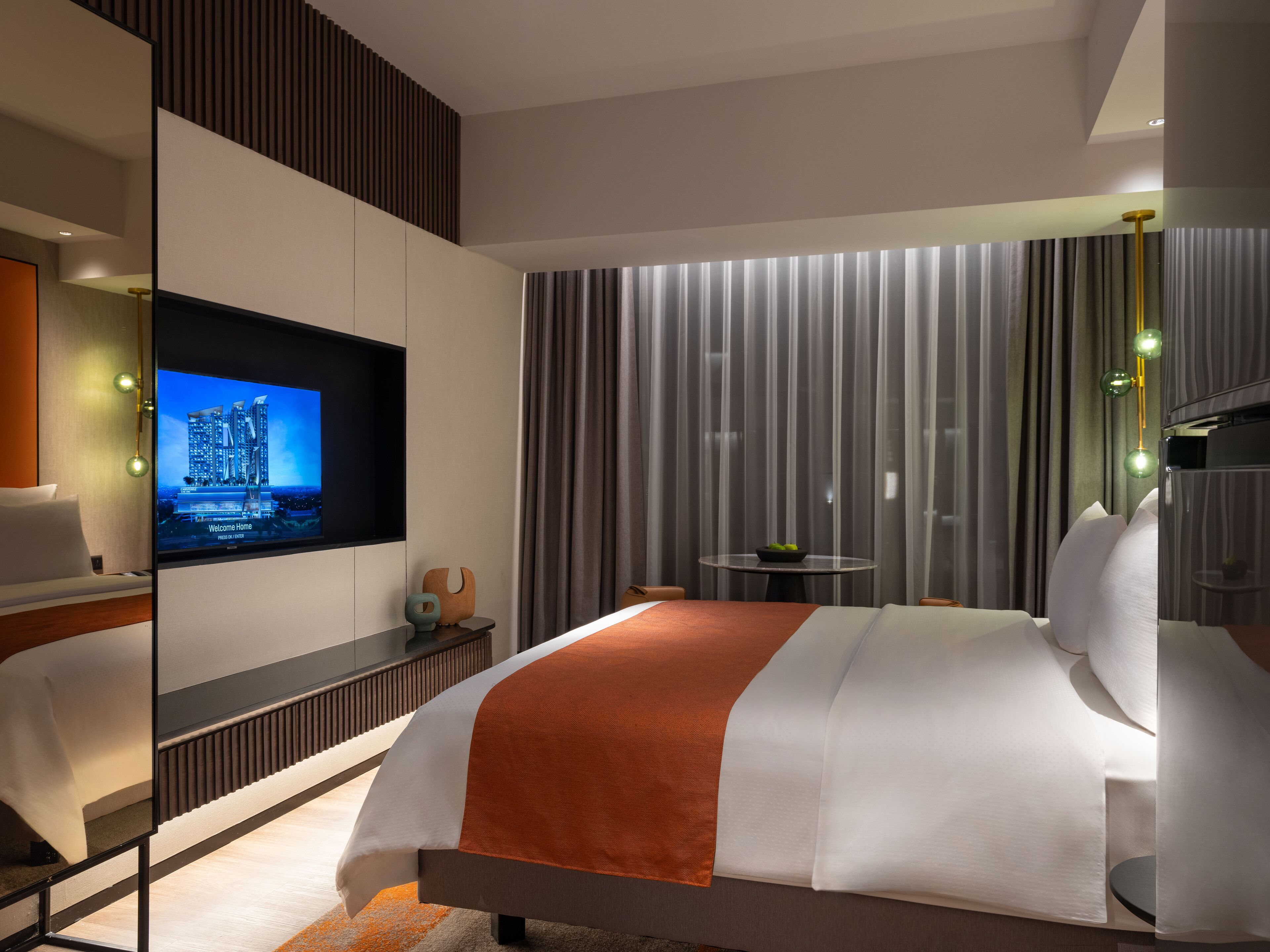 A modern hotel room with a large bed, a flat-screen TV, a small table with a bowl, and a mirror reflecting the room.