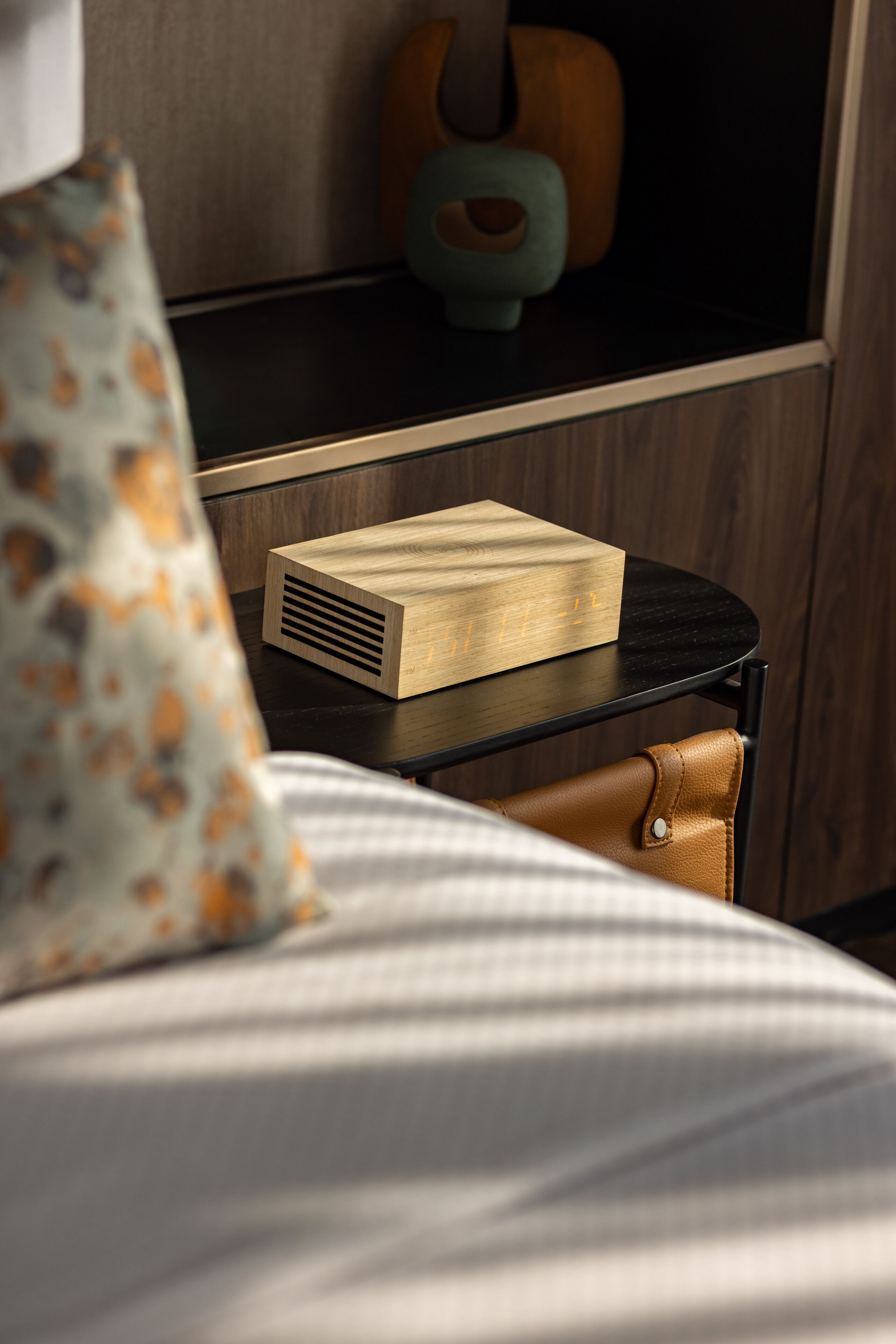 Een netjes opgemaakte slaapkamer met een bed, een nachtkastje met een houten doos, een gestreept kussen en een kleine bruine lederen tas.