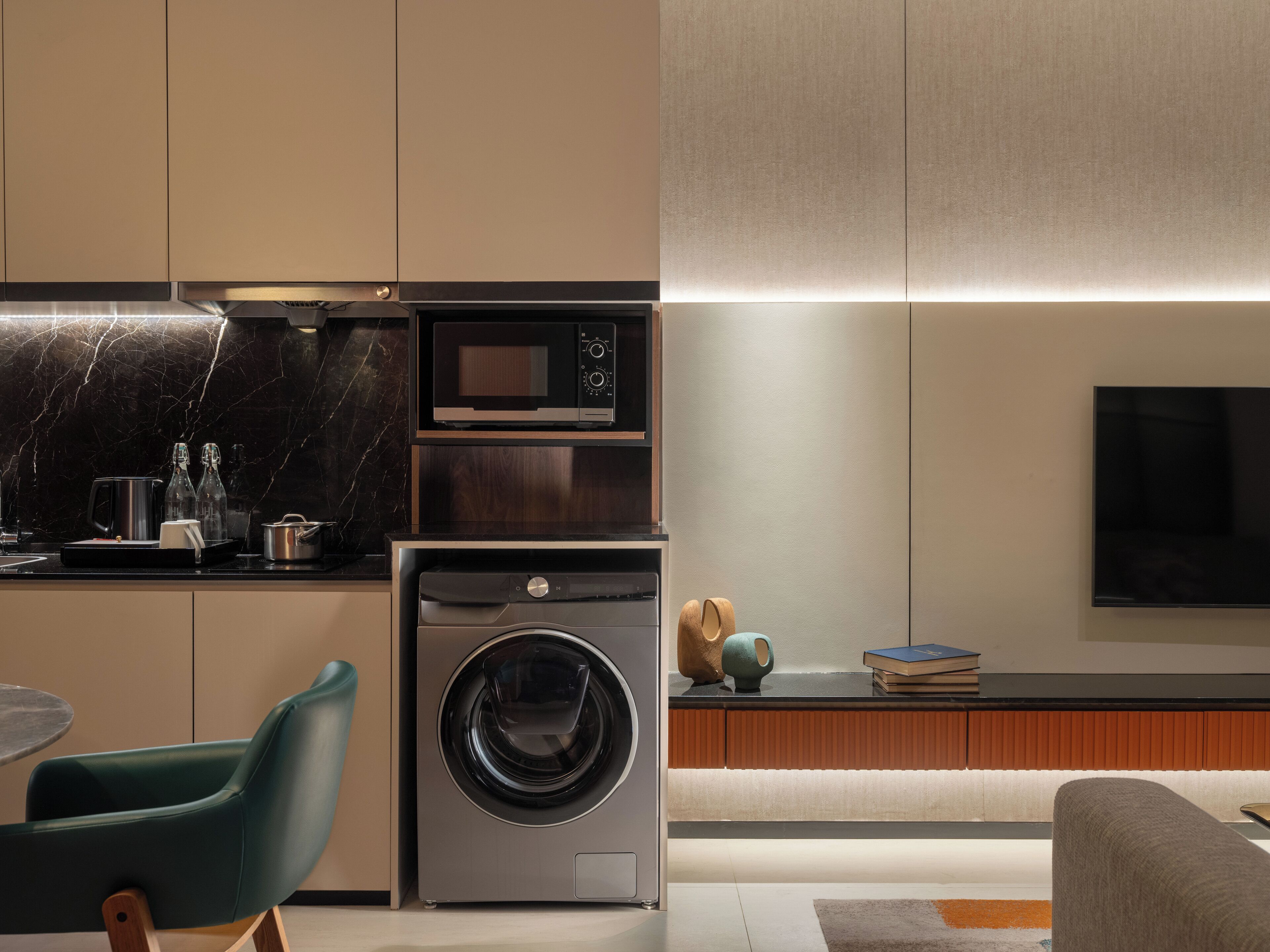 A modern kitchen with a built-in oven, microwave, and washing machine. There is a dining area with a table and chairs, and a sofa in the living room.