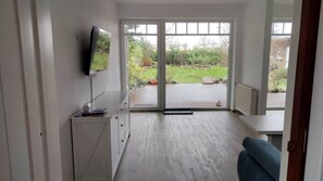 A modern room with a television on the wall, a small desk, and a view of a garden through large sliding glass doors.