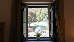 Uma janela com cortinas abertas, um vaso com planta no parapeito e vista para área externa com árvores e uma rede.
