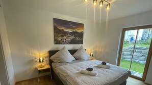 A bedroom with a large bed, two bedside tables with lamps, a painting of a landscape, and a view of a garden through a sliding door.