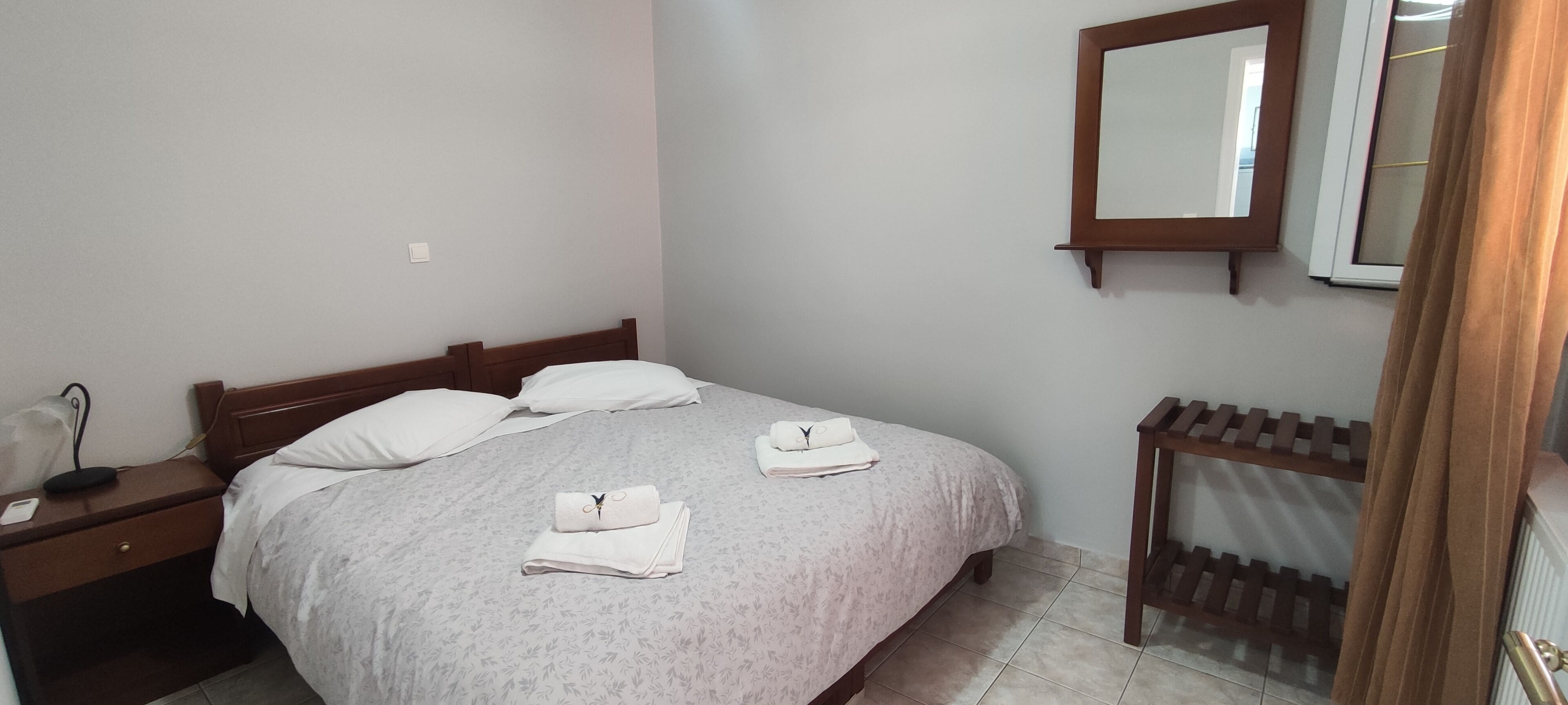 A neatly made bed with a floral bedspread, two pillows, and two folded towels. A wooden nightstand with a phone and a lamp is beside the bed. A mirror hangs on the wall above the bed.