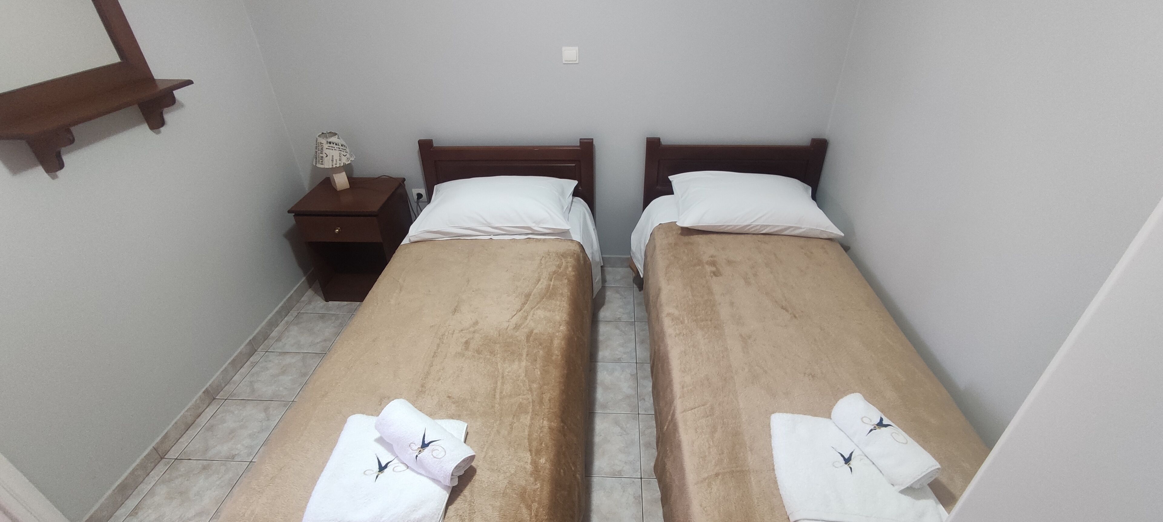 Een kleine, smalle kamer met twee eenpersoonsbedden, elk met een houten hoofdbord en witte beddengoed. Er is een klein houten nachtkastje met een lamp en een klein plankje erboven. De kamer heeft een betegelde vloer en lichtgrijze muren.