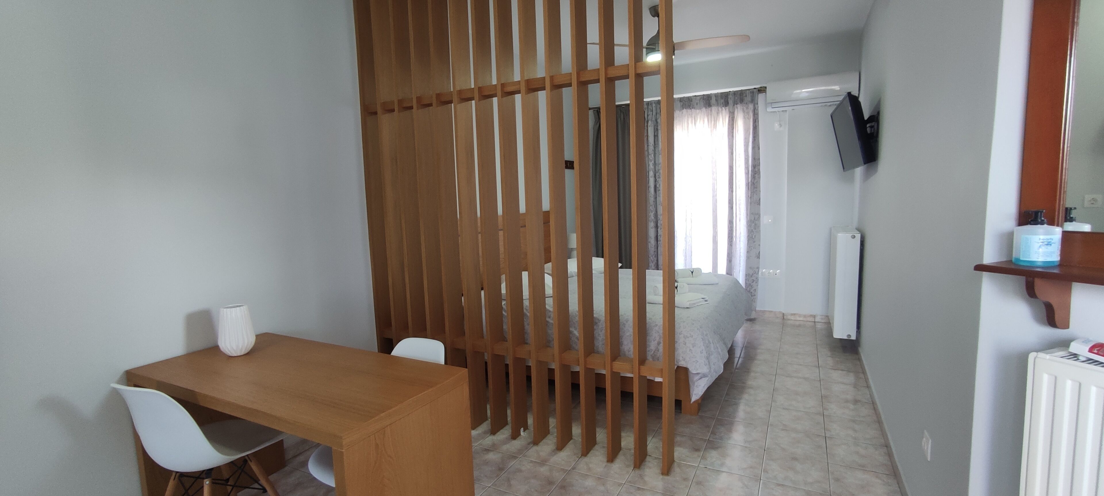 A room with a wooden desk, white chairs, a bed with a white duvet, a wooden partition, a television on the wall, and a radiator.