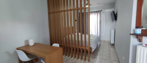A room with a wooden desk, white chairs, a bed with a white duvet, a wooden partition, a television on the wall, and a radiator.