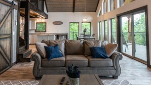 A spacious living area with a large leather sofa, a coffee table with a vase, and a view of the kitchen and outdoor patio. - The Wanderlust Cabin (Ponca)