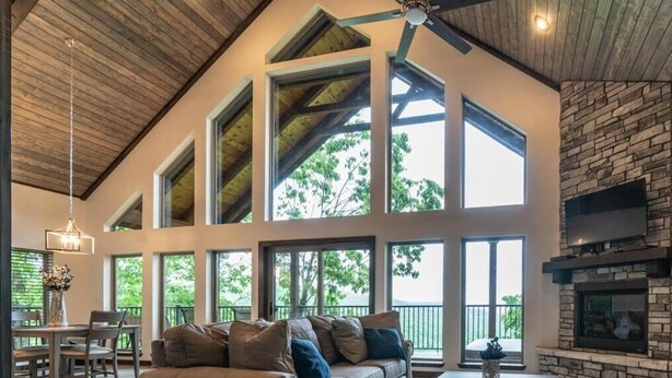 A spacious living room with a high ceiling, large windows, a stone fireplace, and a cozy seating area. - The Wanderlust Cabin (Ponca)