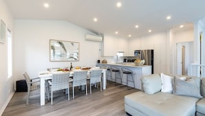 A modern kitchen with white cabinetry, a central island, and a dining area with wicker chairs. - Beach Haven, 3a Messines Street - 4 Minute walk to the beach, WiFi , Air Con (Shoal Bay)