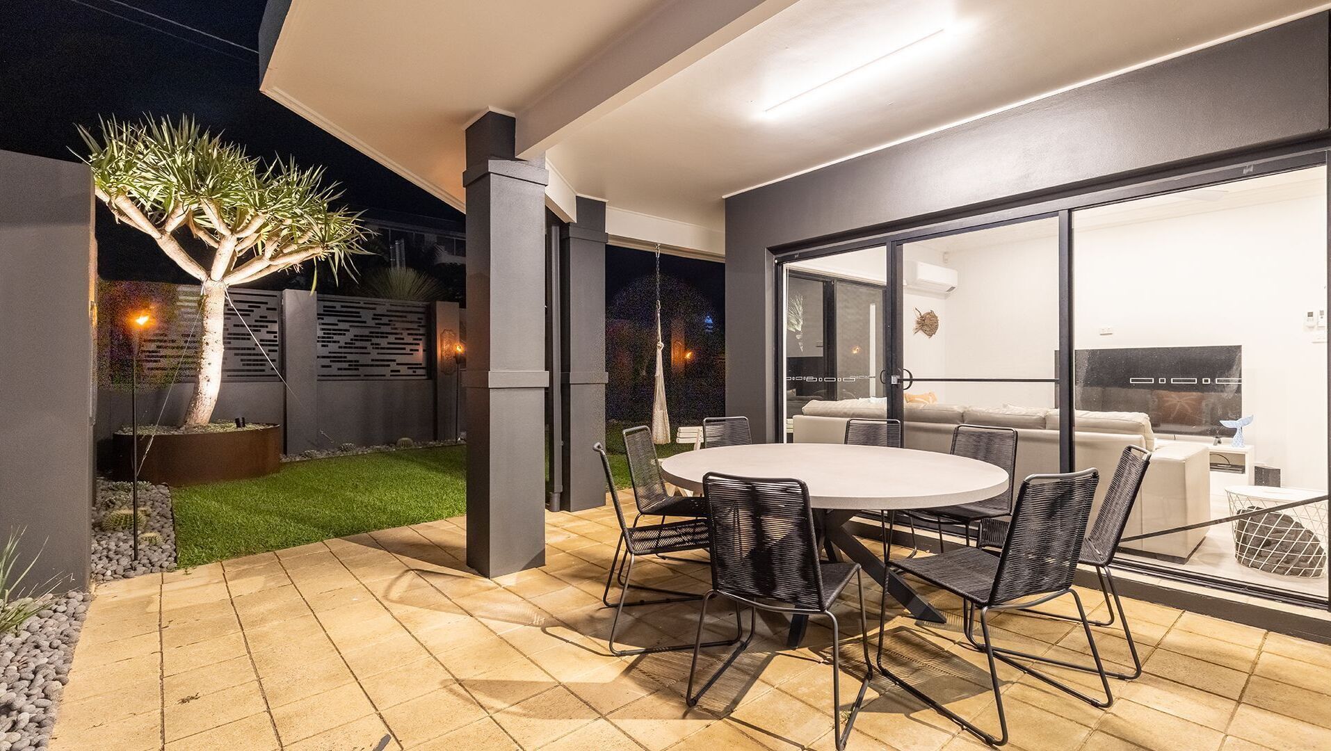 Une terrasse extérieure moderne, dotée d’une table ronde et de chaises, d’un arbre en pot et d’une porte vitrée donnant sur un espace de vie intérieur.