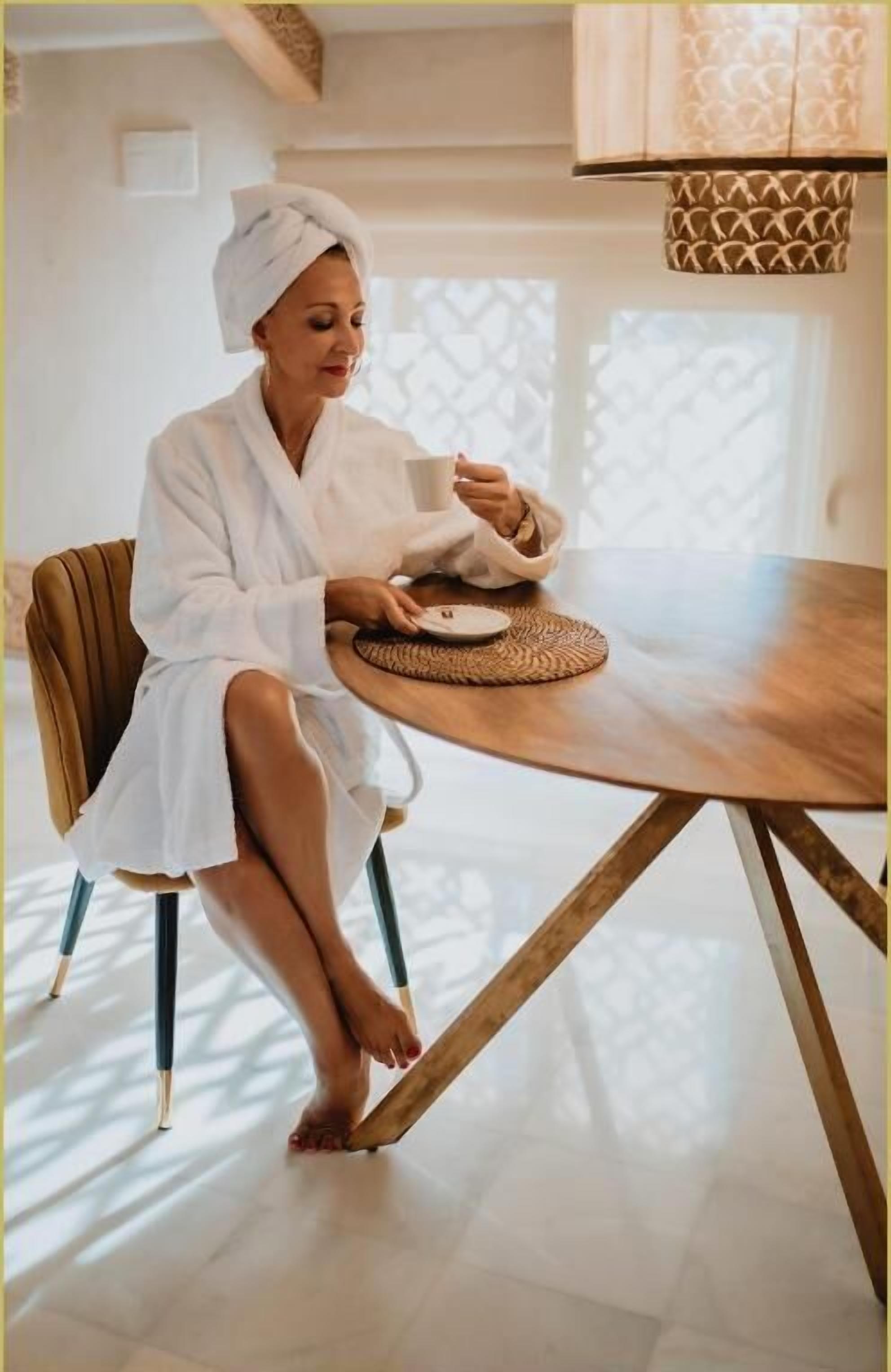 A person in a white bathrobe sitting at a wooden table with a woven placemat, in a room with a modern design and a patterned window.