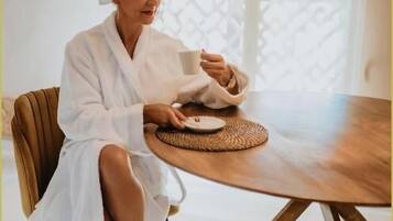 A person in a white bathrobe sitting at a wooden table with a woven placemat, in a room with a modern design and a patterned window.