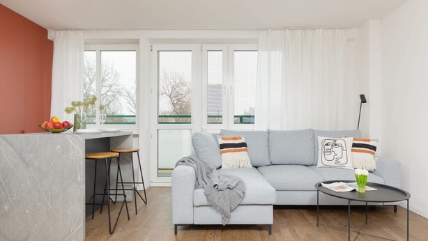 A modern living room with a grey sofa, two bar stools, a small round table, and a kitchen area with a marble countertop.