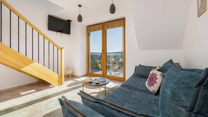 Ein modernes Wohnzimmer mit einer Holztreppe, einem großen Fenster mit Blick, einem blauen Sofa und einem kleinen runden Tisch mit einem Buch und einer Tasse.