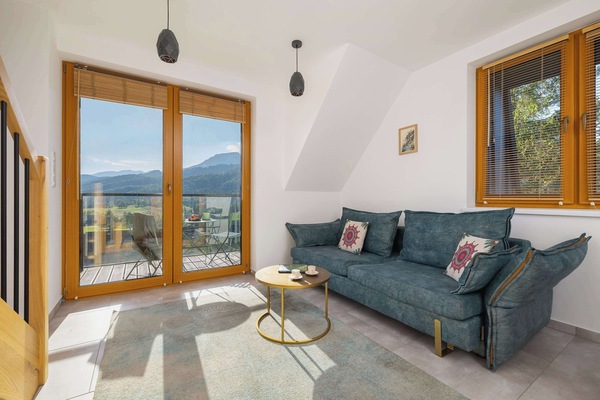 A bright living room with a comfortable sofa set and large windows opening to a balcony with mountain views.