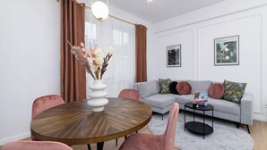 A modern living room with a round wooden table, pink chairs, a grey sofa with patterned cushions, and two framed botanical prints on the wall. - Elegant Apartment | Close to Market | Place to Work (Kraków)