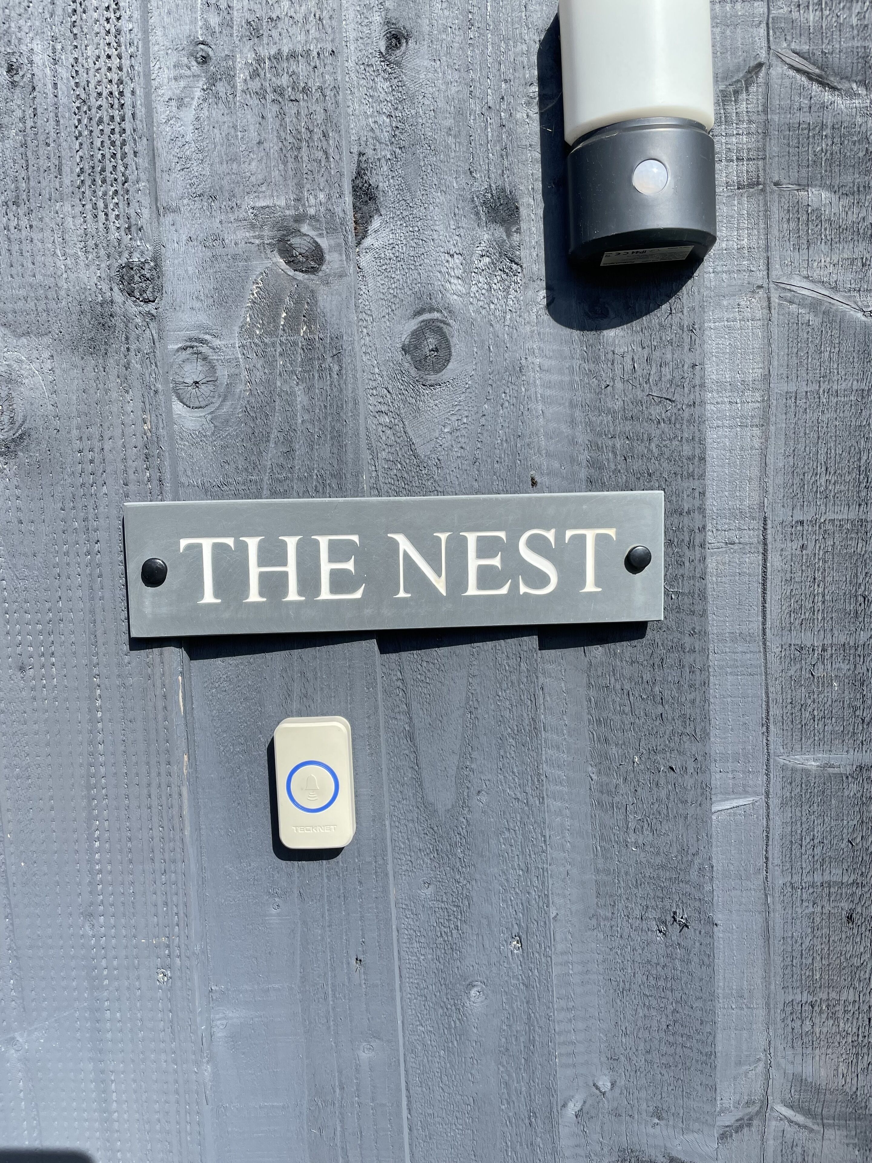 Um letreiro com a inscrição 'THE NEST' em uma superfície de madeira texturizada, um pequeno dispositivo com um círculo azul e um objeto cilíndrico.