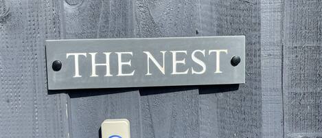A sign labeled "THE NEST" on a textured wooden surface, a small device with a blue circle, and a cylindrical object.