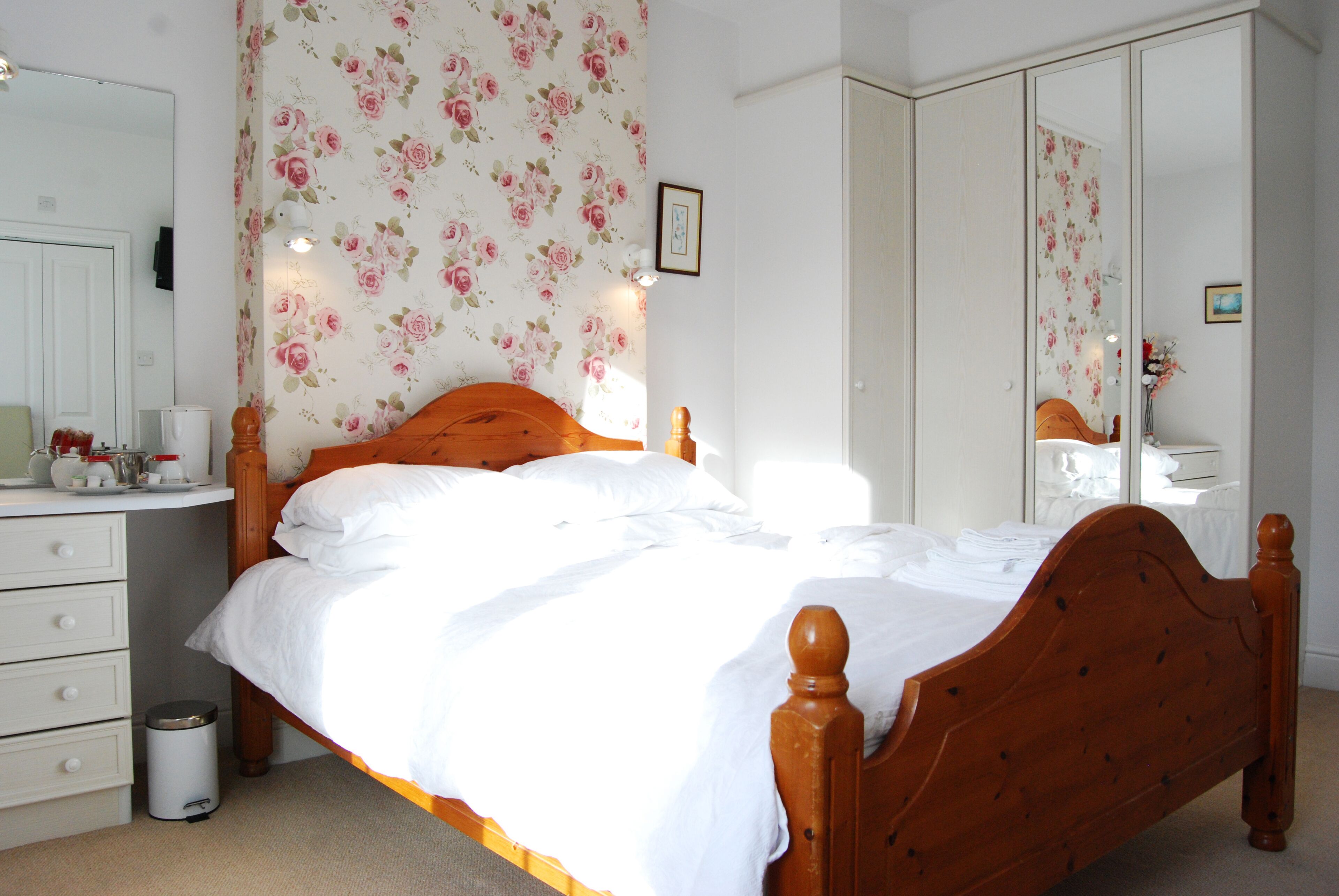 A bedroom with a wooden bed, white bedding, a dresser, a mirror, and floral wallpaper.