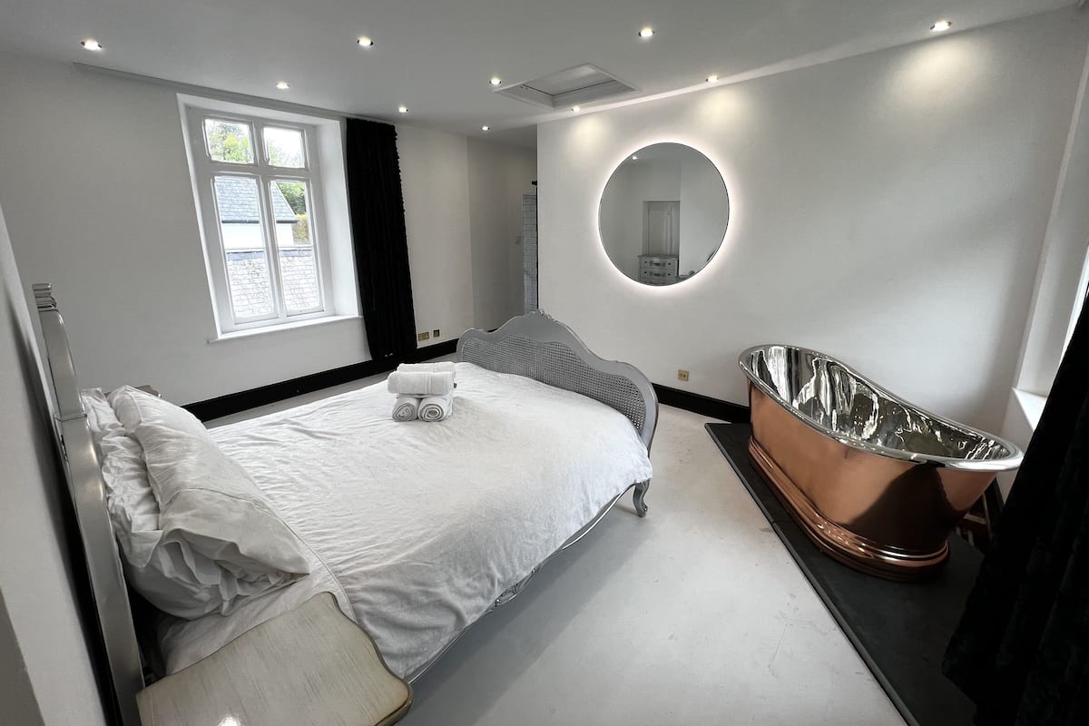 A modern bedroom with a bed, a round mirror, and a freestanding bathtub.