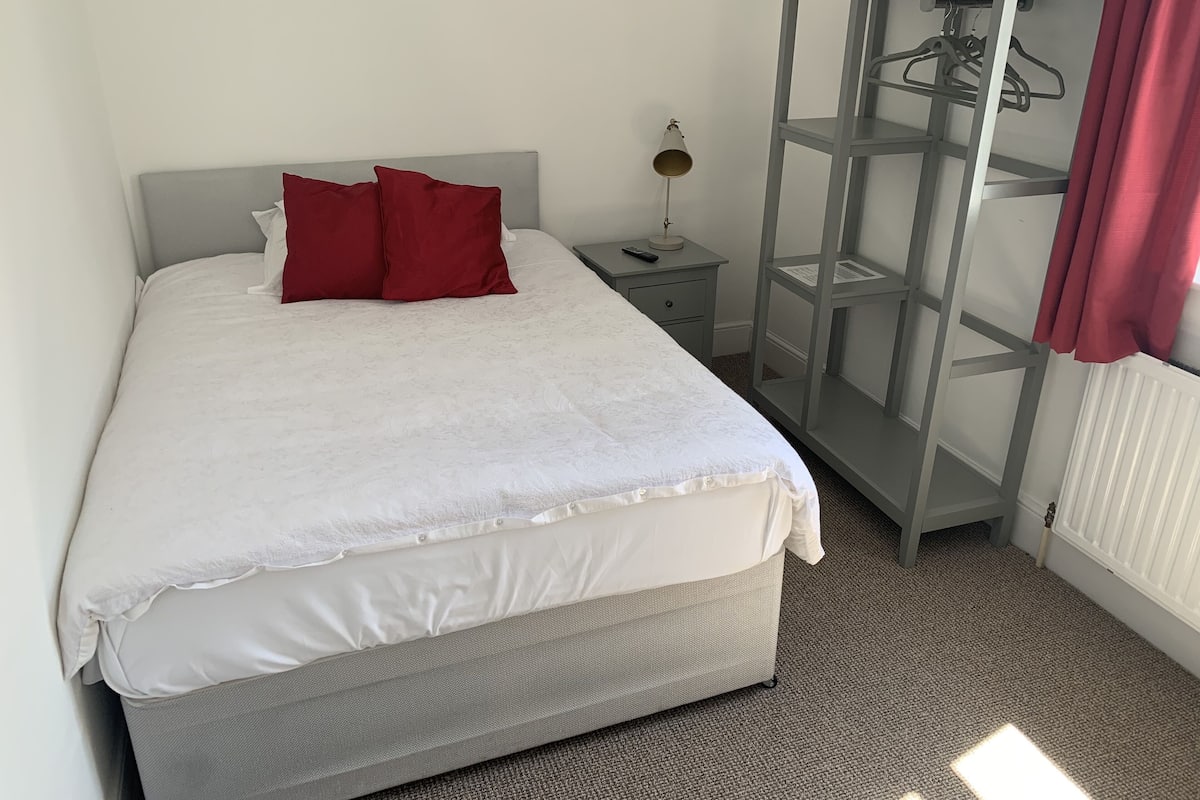 A neatly made bed with white and red bedding, a grey headboard, a bedside table with a lamp, and a grey wardrobe with a mirror.
