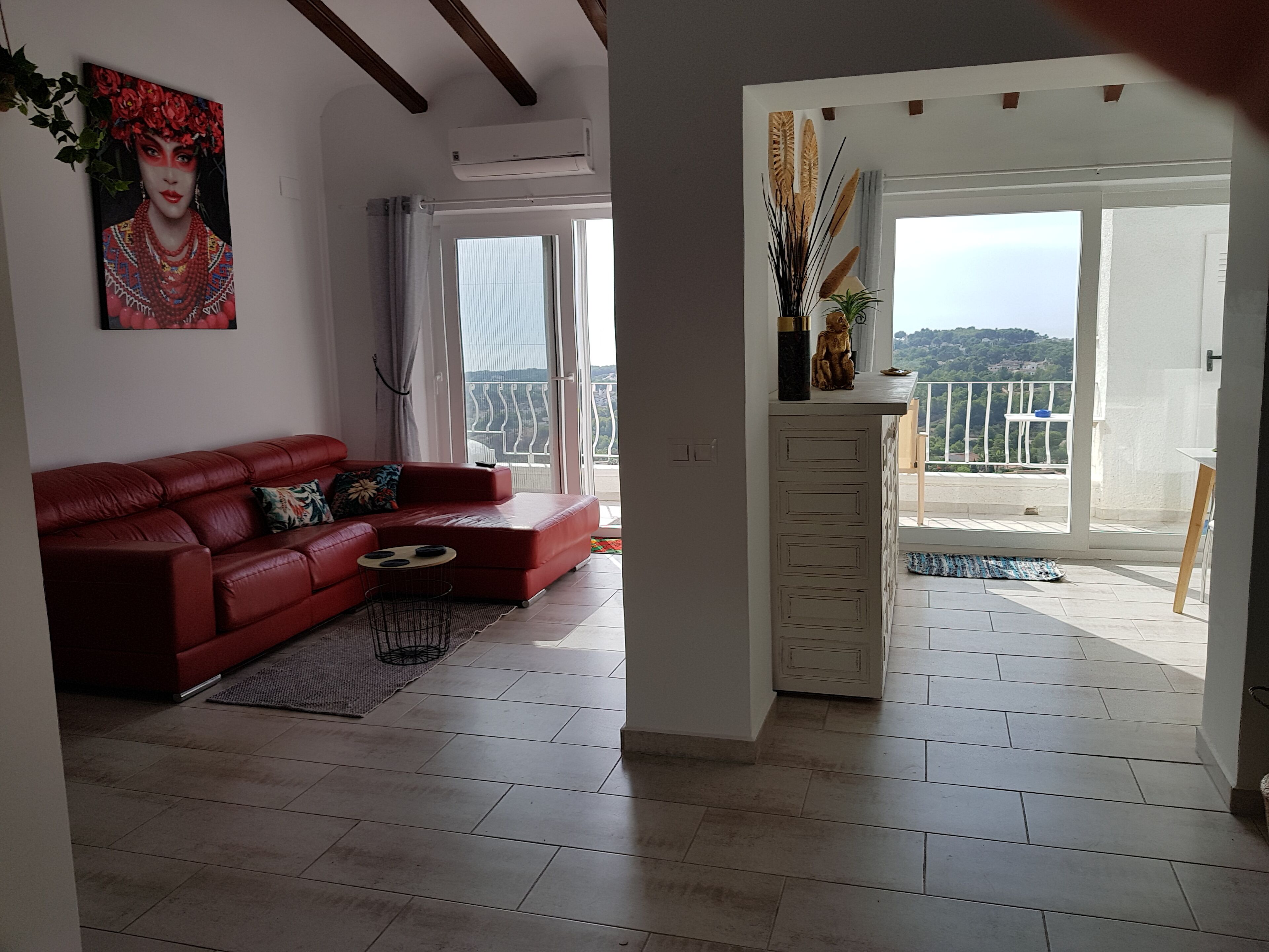 A living room with a red sofa, a small coffee table, and a painting on the wall. There is a balcony with a view of the hills.