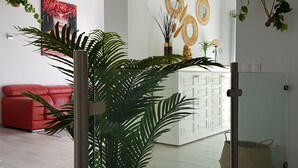 Um interior moderno com um sofá vermelho, uma planta grande em vaso e uma área de cozinha com armários brancos e espelhos decorativos.