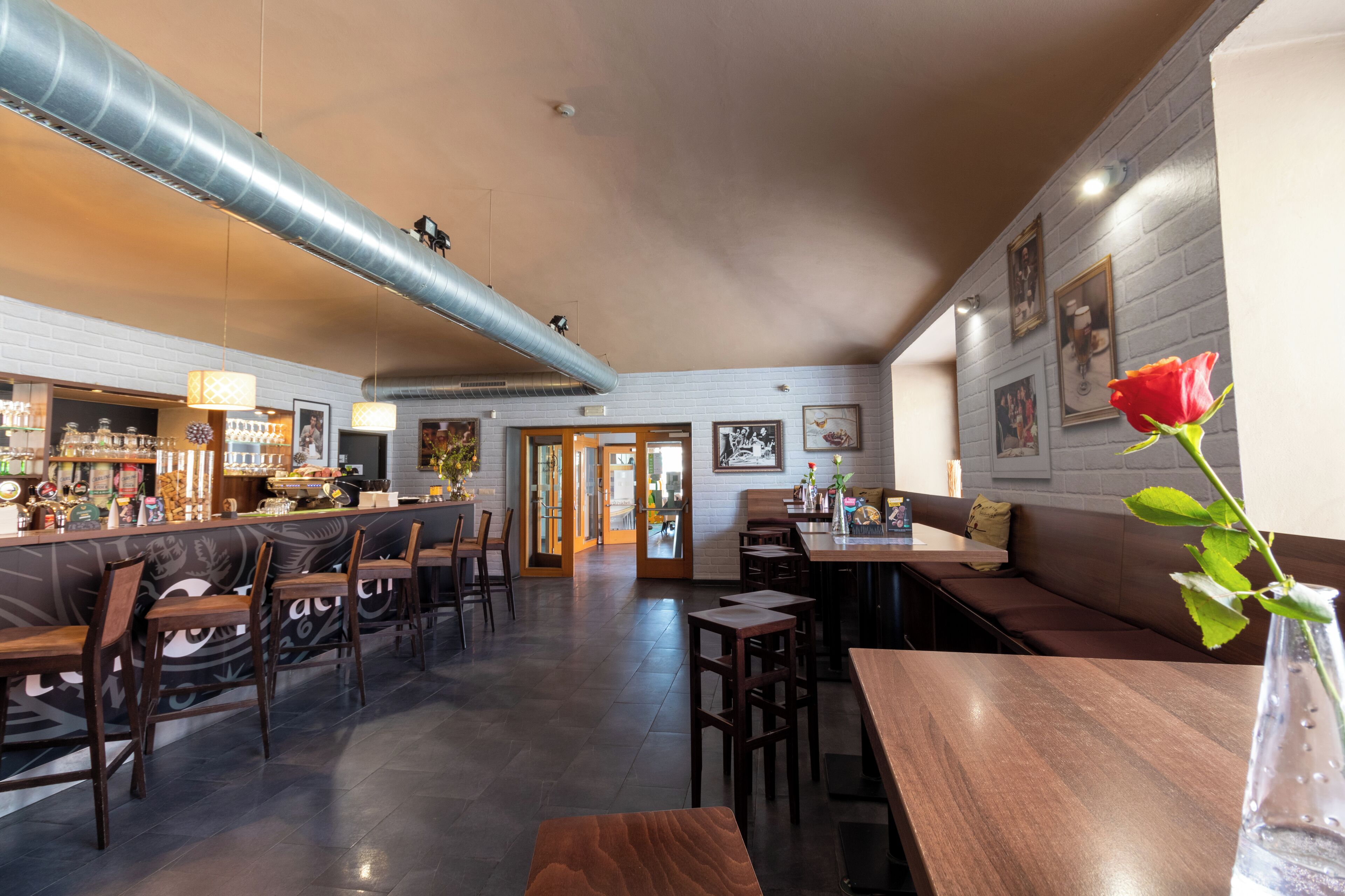 A spacious bar area with wooden furniture, a large ventilation duct, and a red rose in a vase.