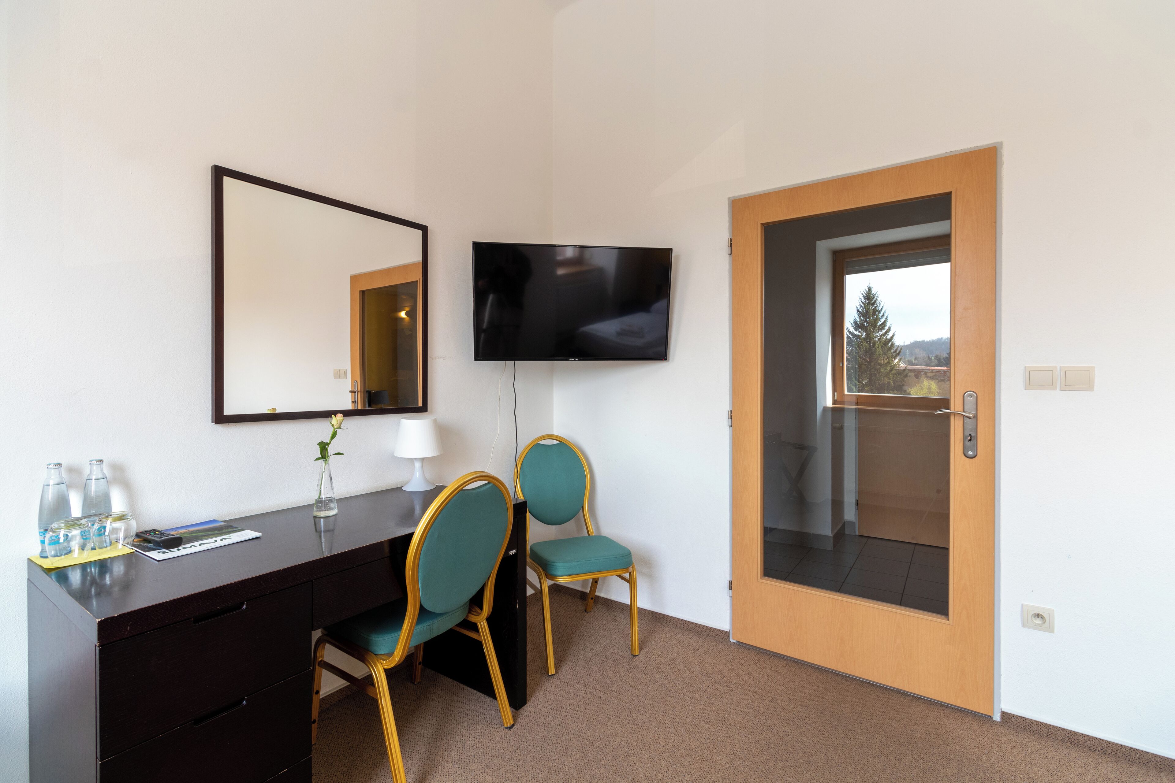 A hotel room with a desk, two chairs, a TV, a mirror, and a door leading to a balcony.