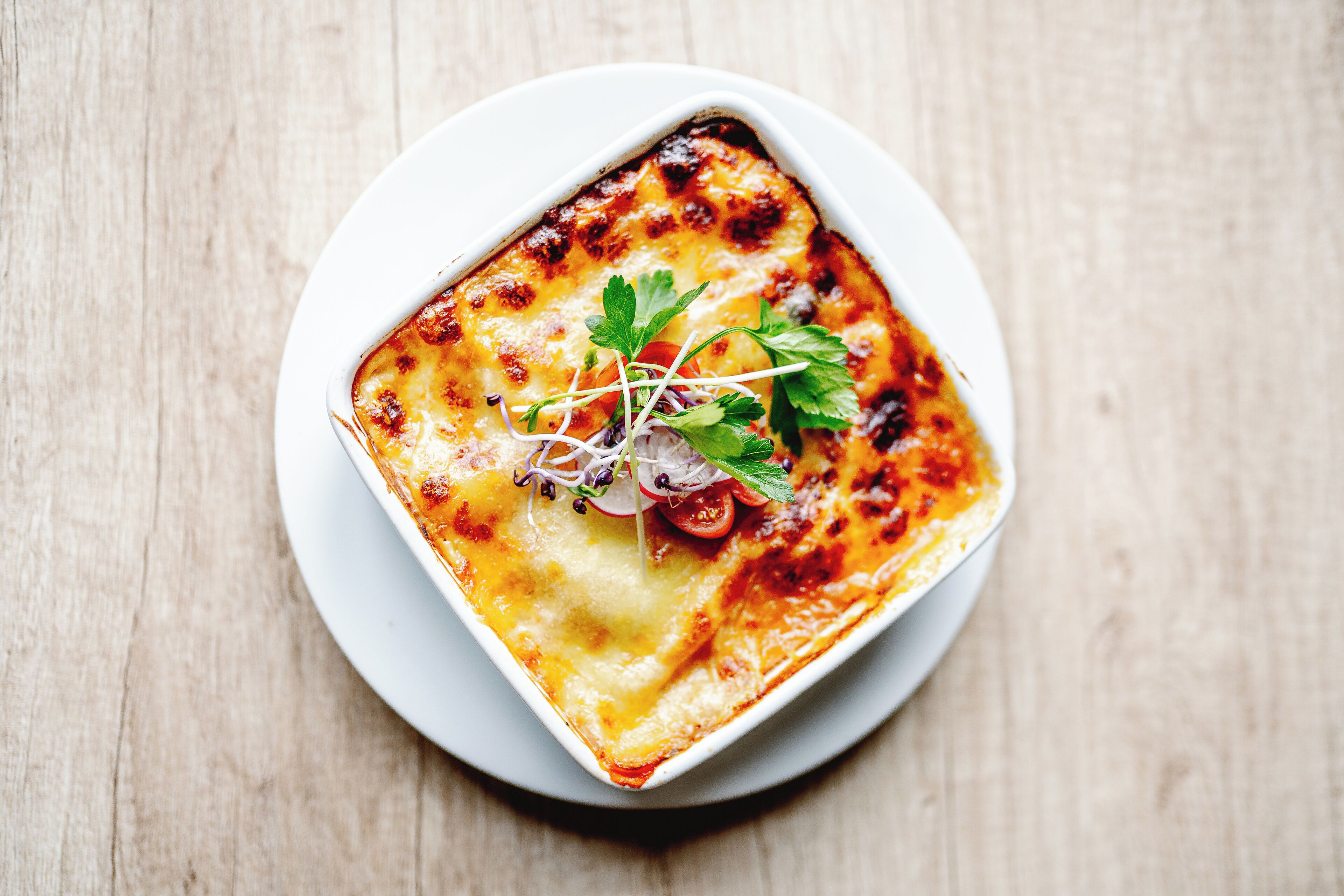 Un piatto quadrato e bianco con una copertura di formaggio gratinato, guarnito con microgreens e fettine sottili di ravanello, su un piatto bianco.