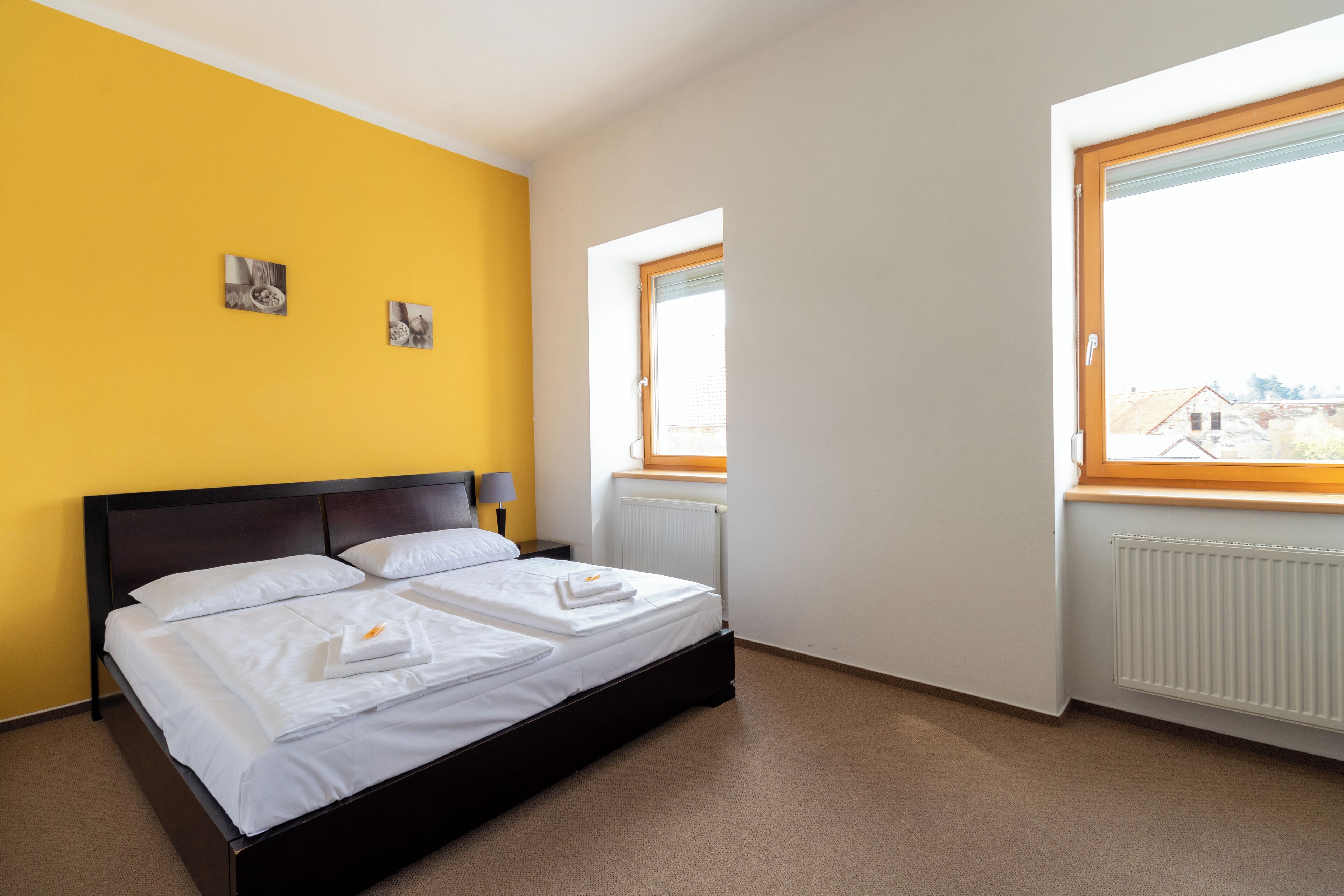A bedroom with a bed, two bedside tables, a window, and a view of buildings outside.